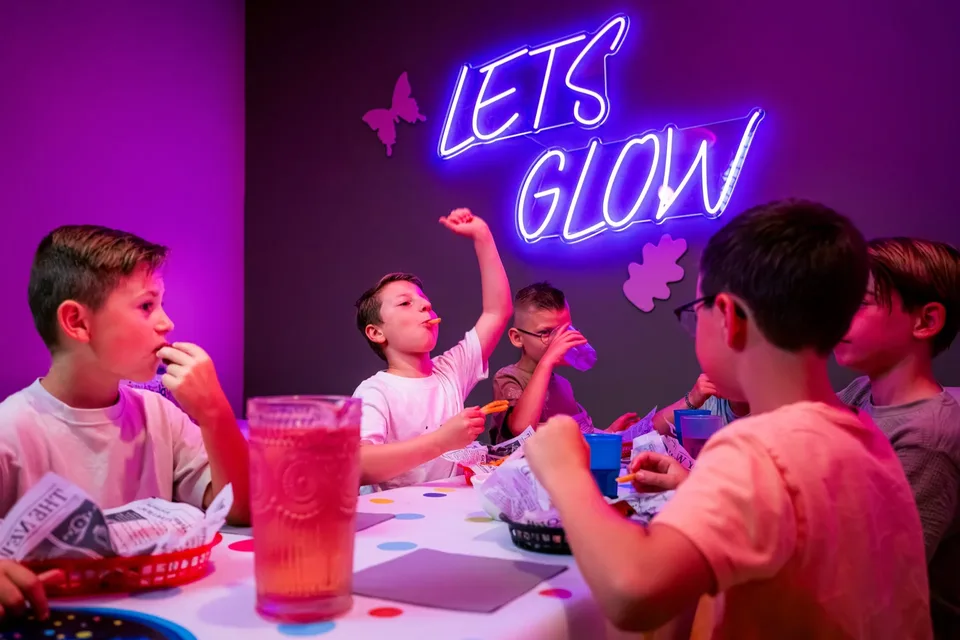 Kinderen eten friet en drinken ranja aan tafel onder de Lets Glow-neon bij een kinderfeestje in Kerkrade